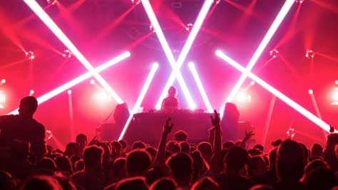 Photograph showing the inside of a nightclub with the DJ taking center stage. The DJ is well lit with red neon lights and a packed dancefloor in front of them. The crowd have their hands up in the air