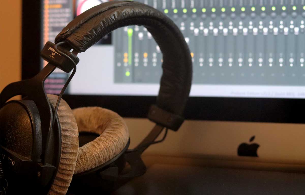 Image of a pair of headphones in the foreground with an Apple Thunderbolt display in the background showing a virtual mixing desk.